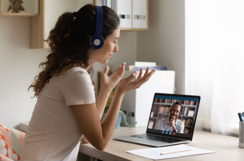 Happy Caucasian young woman look at laptop screen talk chat on video call with African American man husband. Smiling female in headphones have webcam digital virtual communication with friend.